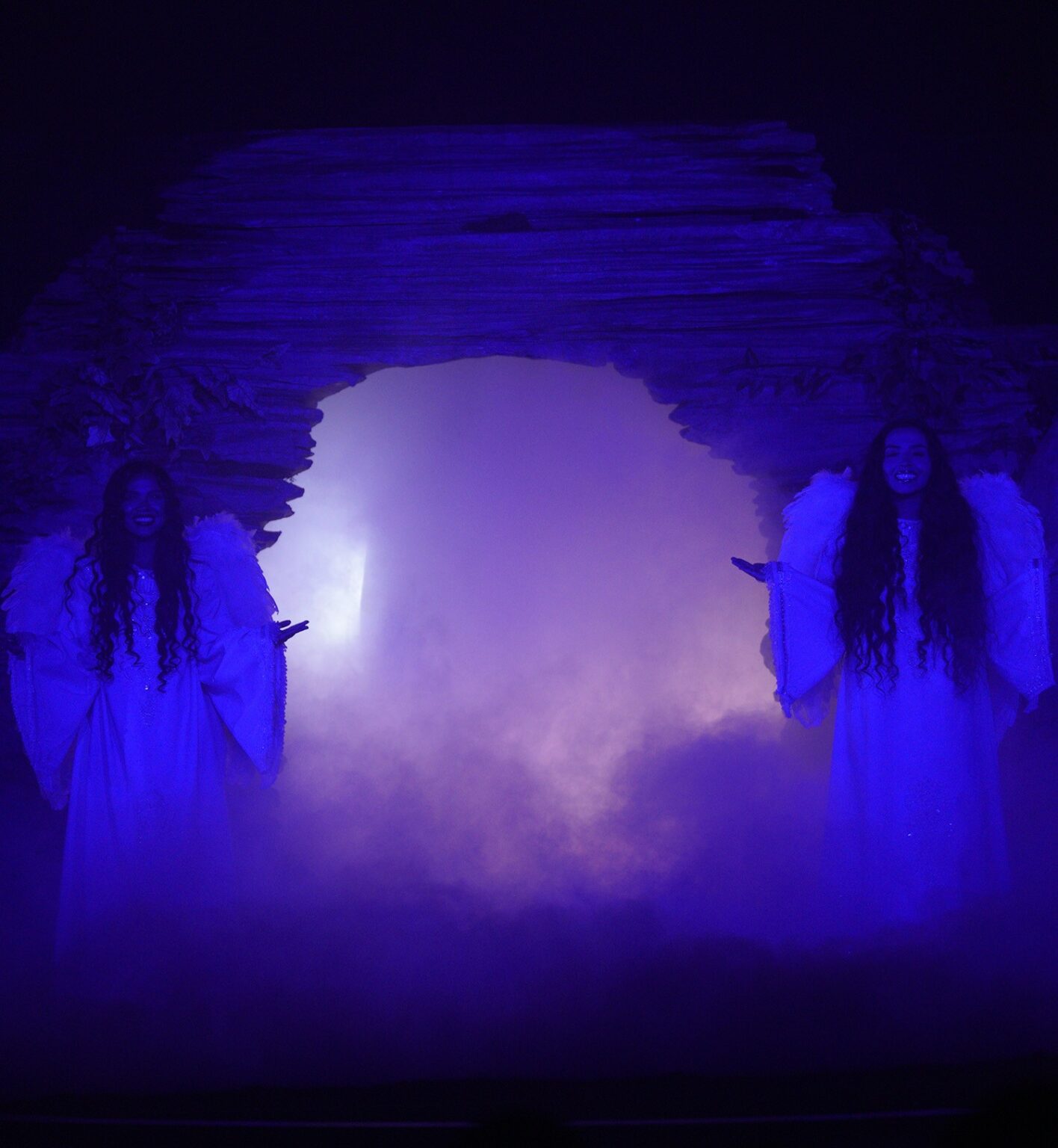 Two people dressed as angles stand on both sides of an archway made of stone. They are bathed in a dark blue light on stage.
