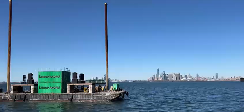 Floating shipping container stage anchored by poles driven into the mud.