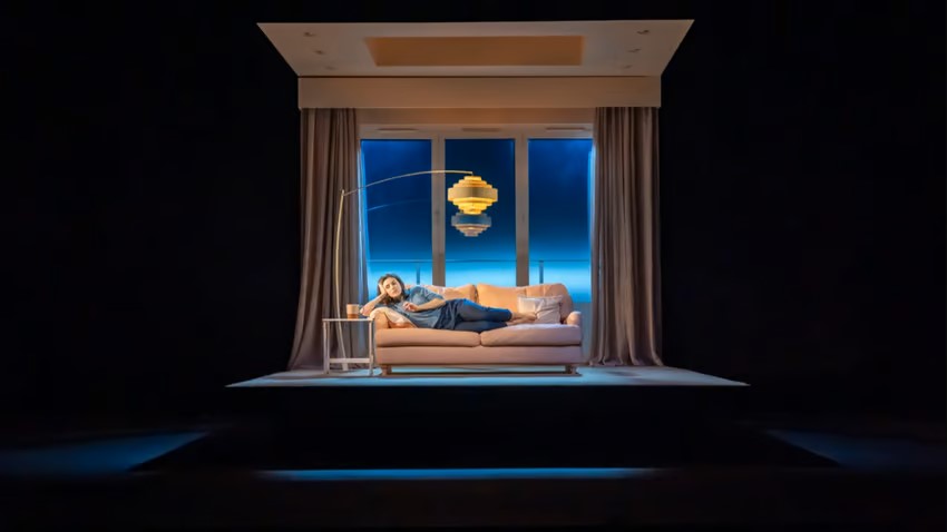 A person lays across a couch on a stage with a large lamp hanging over them. A large picture window is in the background with a blue light coming from it. Johan Persson