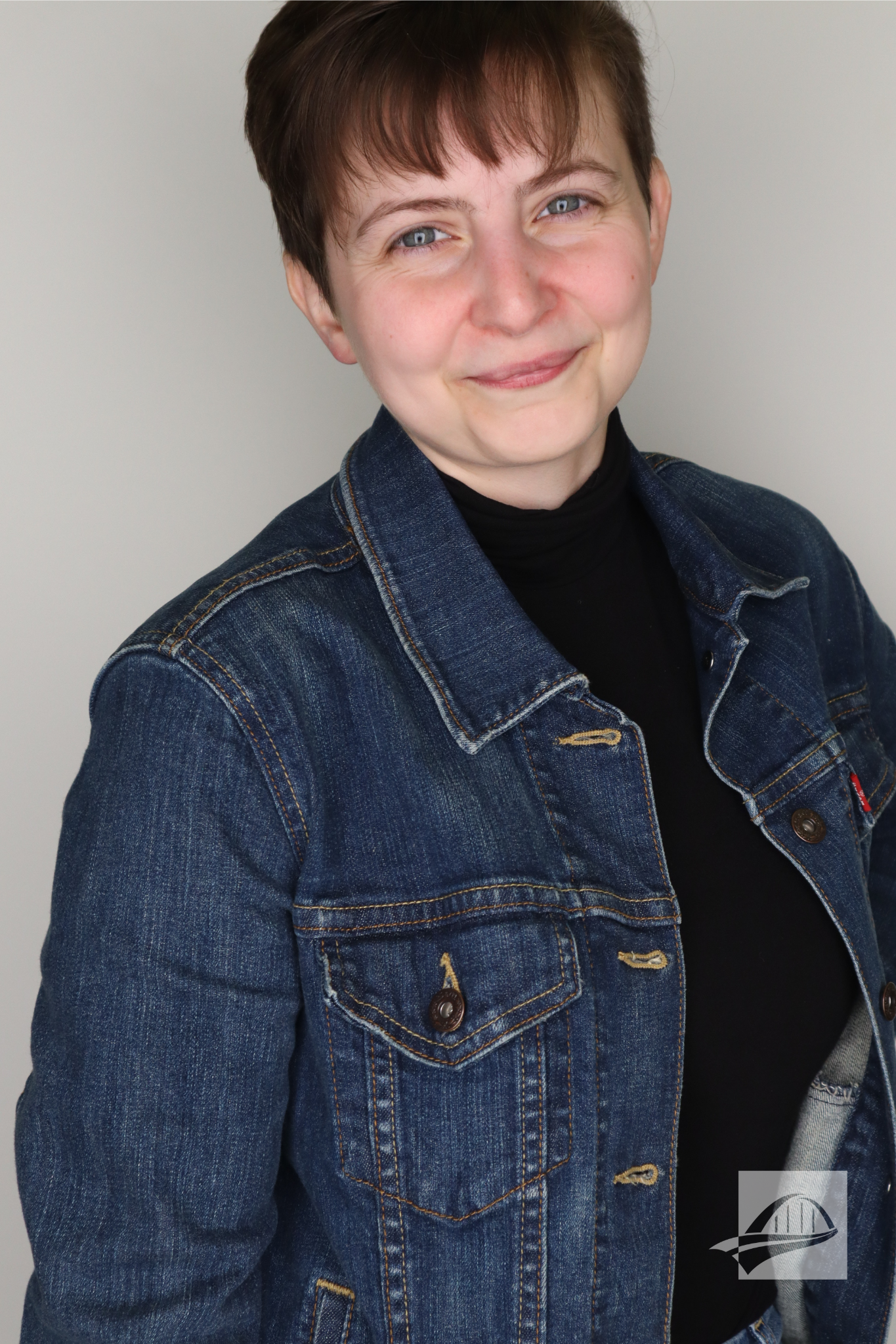 Natalie Houle Headshot. Natalie has short brown hair and is wearing a black turtle neck with a denim jacket over it. She is smiling directly into the camera in front of a gray/white background.
