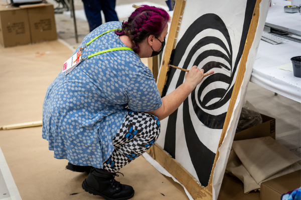 a person in a blue dress squating down and painting a black spiral on a white canvas on the expo floor
