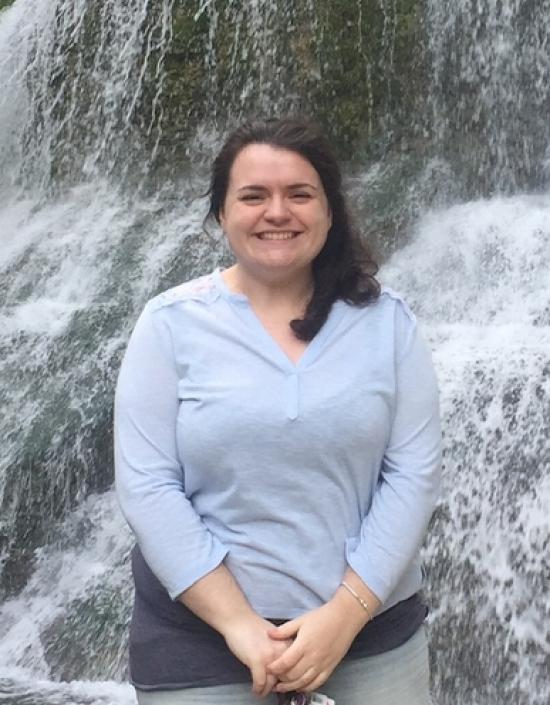 Liz Bilbo stands in front of a waterfall and is wearing jeans, a blue shirt, and has dark brown hair pulled back into a ponytail. She is smiling directly at the camera.